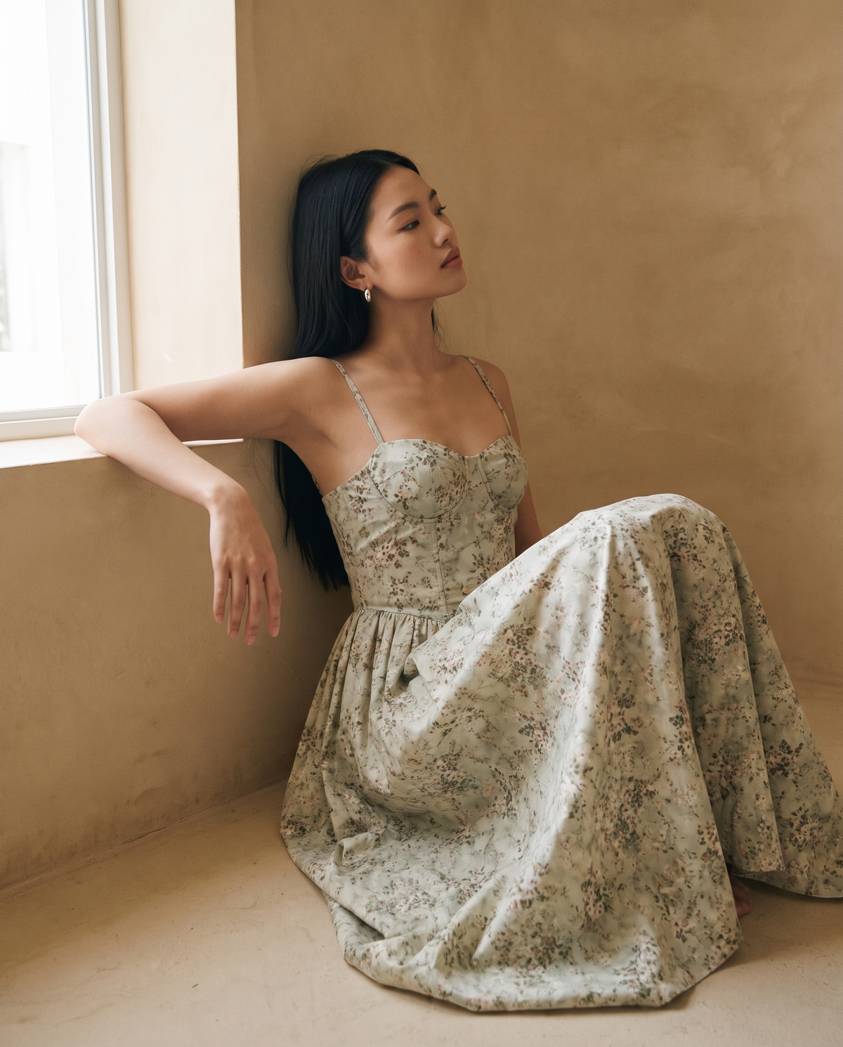 Model seated against a sand plaster wall in the sage floral corset midi dress, soft window light